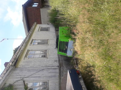 Fotografi av en eksisterende bygning sett fra siden, med en grønn container og høy urt i bakgrunnen.