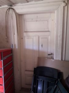 Fotografi av en hvit dør med paneler og et nøkkelhull, sett fra innsiden av et rom. Det er også synlig en rød skuffe til venstre og mørke gjenstander (muligens stolset) foran.