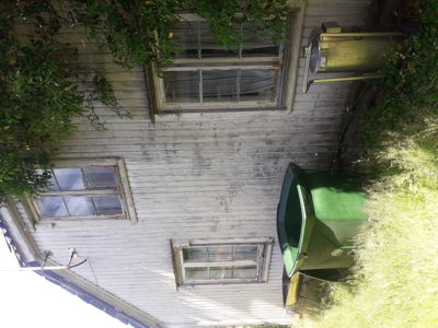 Fotografi av en fasade på et gammelt trehus med vinduer, plankevegg og omgivelser.