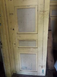 Fotografi av en gammel, gulmalt dør med paneler og synlig slitasje.