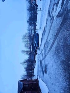 Fotografi av en snødekt vei med parkerte biler og en rødbrun hytte til venstre.