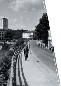 Et svart-hvitt fotografi som viser en gateoppsett med en kirke i bakgrunnen, en person som går på en bro eller gangvei, og syklister på veien.