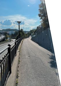 Fotografi av en eksisterende gangvei eller sykkelsti langs en vei med en betongmur og en hekk.