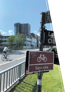 Fotografi av en gate med sykkelskilt, syklist og bygninger i bakgrunnen.