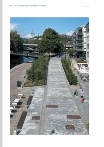 Fotografi av en utendørs gangvei eller promenade med mønstret belgstein, omgitt av trær, en elv og bygninger.