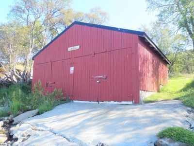 Fotografi av en rød, langhusformet bygning (sannsynligvis en uthus eller skur) sett fra en vinkel som viser både fasade og sidevegg. Bygningen har store dører, er omgitt av trær og har en betongplate foran.