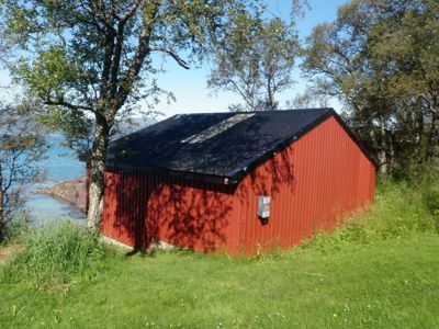 Fotografi av en rød, metallisk bygning (trollbustad eller uthus) med svart tak, plassert i et naturområde med trær og vann i bakgrunnen.