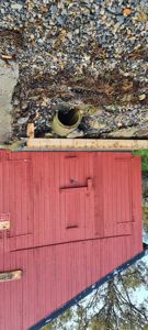 Fotografi av en rød, trepanelert bygning (kanskje en uthus eller skur) sett fra siden. Bildet viser bygningens fasade, en utløpsrør under bygningen, og omgivelsene med grus og vegetasjon.