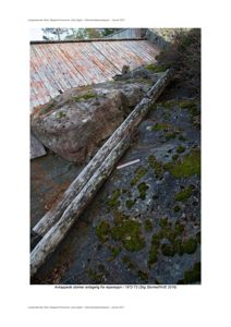 Fotografi av en stor steinblokk og to store tømmerstokker som ligger på bakken ved siden av et bygning med rustet tak.