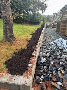 Fotografi av en byggeplass som viser en ny anlagt betongmur, en gravet grøft med jord, og en belte med stein. I bakgrunnen er det synlig en del av en bygning og vegetasjon.