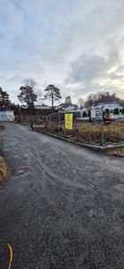 Fotografi av en byggeplass med en gjerde, et gul skilt og en grusvei i forgrunnen.