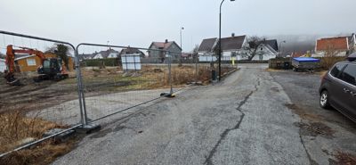 Fotografi av en byggeplass med en grøft, en gressklipper, en bil og nabolagshus i bakgrunnen.