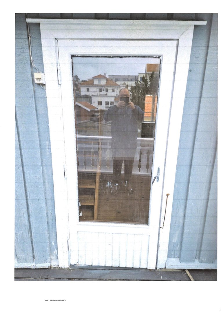 Fotografi av en hvit dør med glassfelt i en blå fasade. Refleksjonen i glasset viser en person som tar bildet.