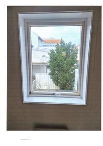 Fotografi av et takvindu sett fra innsiden av et rom. Bildet viser vindusrammen, taket rundt og utsikt gjennom vinduet til nabohus og en stor busk.