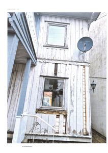 Fotografi av en eksisterende, hvitmalt trebygning sett fra siden. Bygningen har et utslitt fasade med en satellittantenne og en lampe.