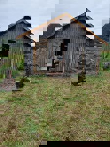 Fotografi av en gammel, gråtrekonstruert uthus eller skur med åpen dør, plassert på en grøntbeveget tomt med fjell i bakgrunnen.