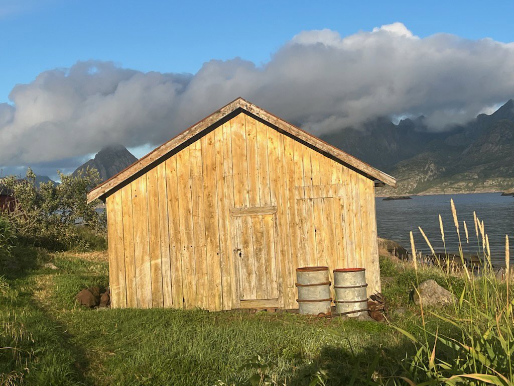 Fotografi av en liten, gammel trehytte eller uthus plassert i en naturskjønn omgivelse med fjell og vann i bakgrunnen.
