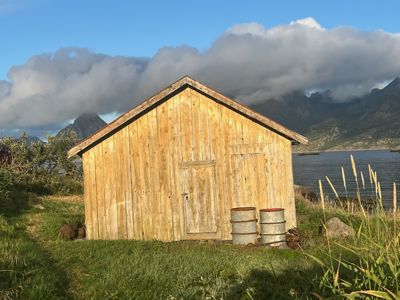 Fotografi av en liten, gammel trehytte eller uthus plassert i en naturskjønn omgivelse med fjell og vann i bakgrunnen.