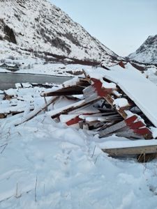 Fotografi av en nedrevet bygning med snødekte takplater og konstruksjonselementer i forgrunnen, med fjell og vann i bakgrunnen.