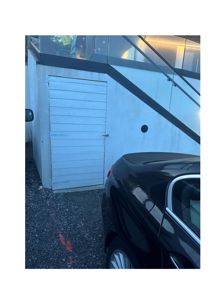 Fotografi av en hvit, panelert dør (sannsynligvis til en kjeller eller garasje) i en bygning med glassfasade og trapp oppover. En svart bil er parkert foran.