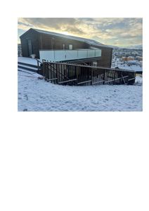 Fotografi av en moderne, mørk trehytte med snø på bakken og en byutsikt i bakgrunnen.