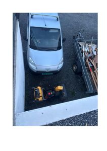 Fotografi av en hvit varebil, en gressklipper og en henger med byggavfall parkert på et grusareal ved siden av en betongmur.