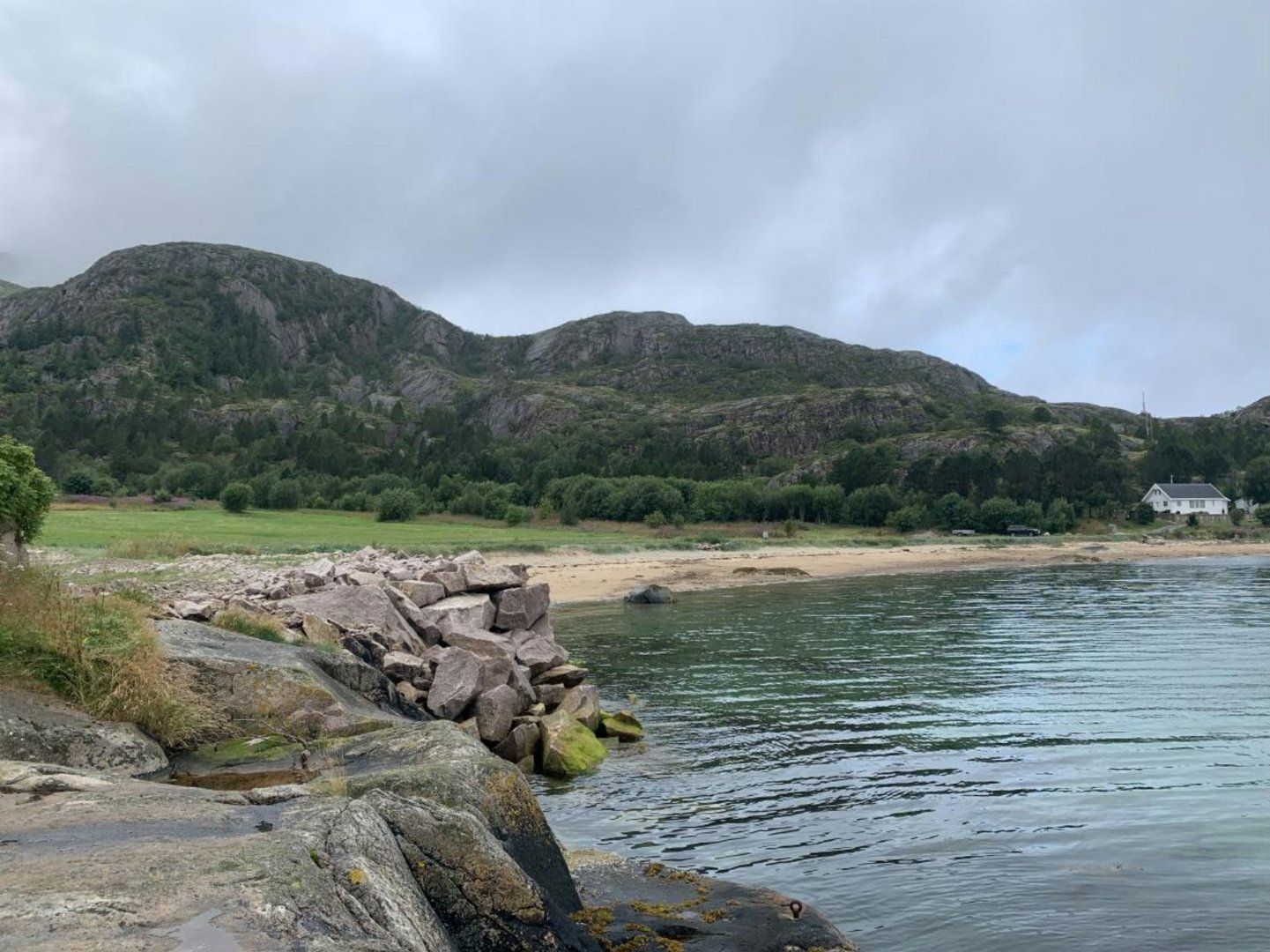 Fotografi av en kyststrøk med fjell, strand og vann. Bildet viser et eksisterende landskap og en bygning i bakgrunnen.