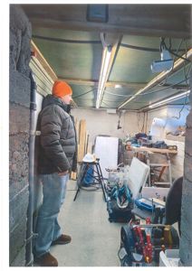 Fotografi av en mann som står i et underjordisk rom (keller eller kjeller) under bygging eller renovering. Mannen er kledd i vinterklær og ser ut til å inspisere rommet. Rommet inneholder byggematerialer, verktøy og utstyr.