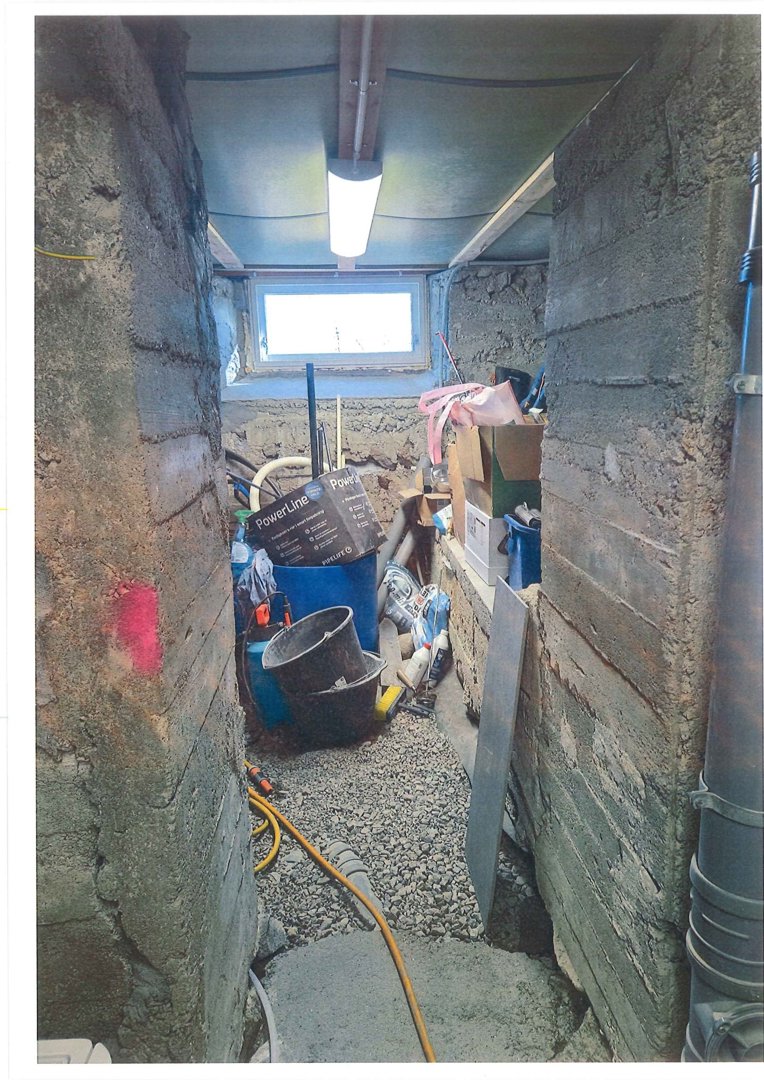 Fotografi av en underjordisk eller halvunderjordisk gang i en bygning under ombygging eller vedlikehold. Bildet viser råbetongvegger, gulv med grus, rør og byggemateriale.