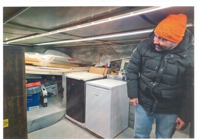 Fotografi av en mann i en oransje lue og mørk jakke, som står i et rom med utstyr og materialer i bakgrunnen.