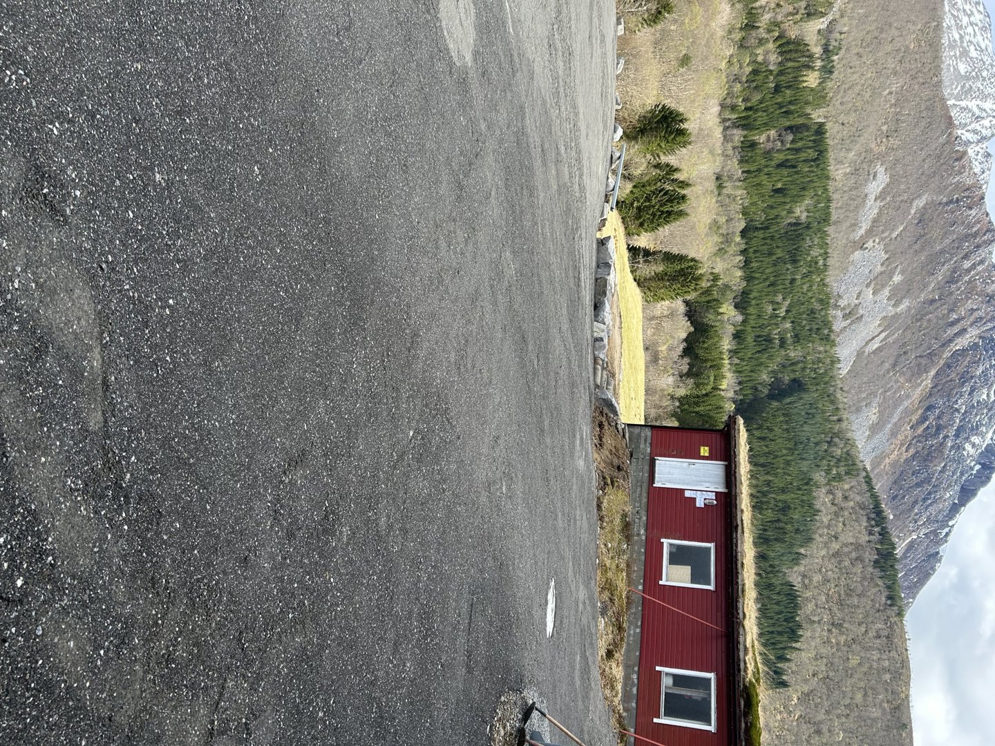 Fotografi av en liten rød bygning med grønt tak (sannsynligvis et jordtak) sett fra en asfaltert overflate. Bakgrunnen viser et fjellandskap med skog og fjell.