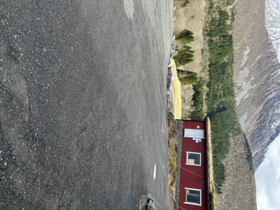 Fotografi av en liten rød bygning med grønt tak (sannsynligvis et jordtak) sett fra en asfaltert overflate. Bakgrunnen viser et fjellandskap med skog og fjell.