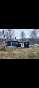 Fotografi av en mørk, trolig trebygning (hus eller hytte) sett fra siden, omgitt av bartrær og grøftete terreng.