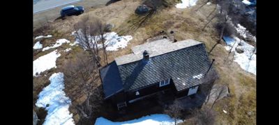 Fotografi av en mørk, trolig trebygning sett fra luften (dronebilde). Bygningen har et skifer- eller taktegltekket tak og er plassert på et terreng med rester av snø.