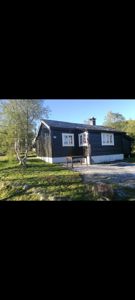 Fotografi av en mørk, trolig svart, hytte eller liten bygning sett fra siden. Bygningen har hvite vindusrammer, en hvit sokkel og en røykledning. Den står på en grønn, grusete tomt med trær i bakgrunnen.