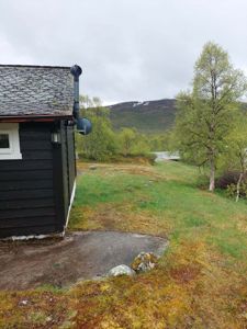 Fotografi av en mørk hytte eller uthus sett fra siden, med en bakgrunn av grønt terreng, trær og fjell.