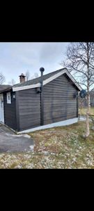 Fotografi av en mørk, trepanelt bygning sett fra siden, med synlig tak, røykledning og omgivende natur.