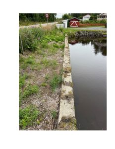 Fotografi av en eksisterende bygning (rød uthus) og omgivelsene, inkludert en vannvei og en vei.