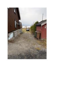 Fotografi av en grusvei mellom to bygninger (en mørk trehus og en rød skur) med en bil i bakgrunnen.