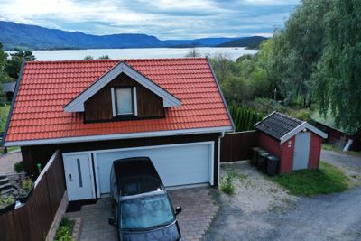 Fotografi av en villa med rød takflis, garasje og en liten uthus ved siden av, sett fra en høyde med utsikt over vann og fjell i bakgrunnen.