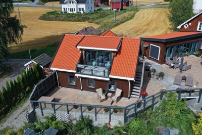 Fotografi av en moderne, mørk trehytte med oransje tak sett fra lufta (dronebilde). Bildet viser bygningens fasade, terrasser, omgivelser og nabolag.