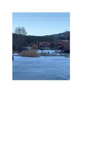 Fotografi av en bygning (eller flere) sett fra et frosset vann eller snødekt område, med bakgrunn av skog og fjell.