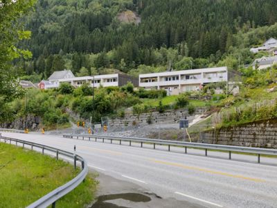 Fotografi av en vei som svinger opp mot en skråning med flere bolighus i bakgrunnen.
