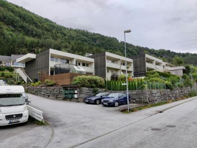 Fotografi av en moderne boligblokk med flere etasjer, parkerte biler foran og en skråning bak.