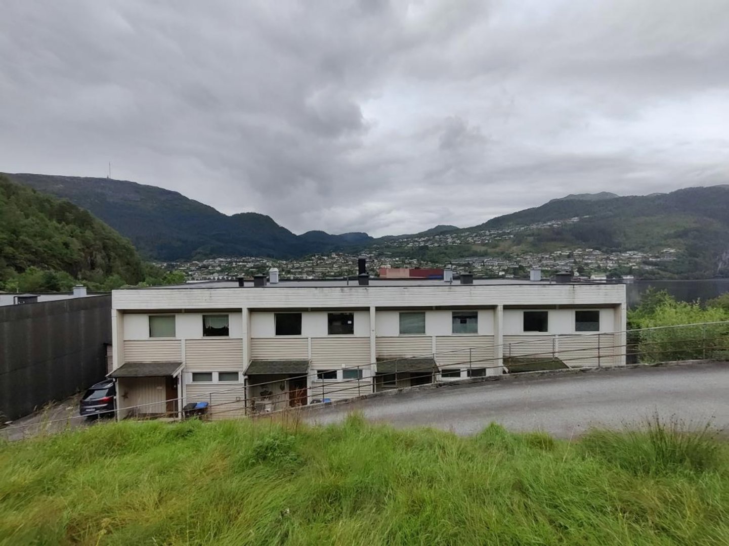 Fotografi av en hvit, toetasje bygning sett fra siden, med bakgrunn av fjell og et byområde.