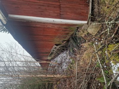 Fotografi av en rød, trepanelt bygning (sannsynligvis en uthus eller garasje) sett fra siden, med omgivende natur og snø på bakken.