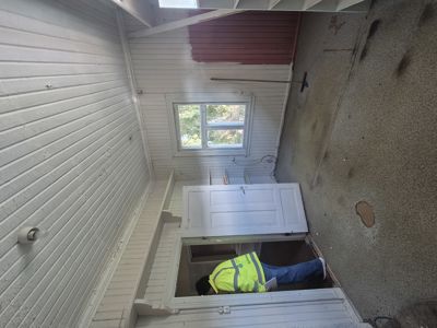 Fotografi av en person i arbeid (gule vest) som utfører reparasjoner eller ombygging inne i et hus, sannsynligvis på gulvet eller ved å fjerne panel.