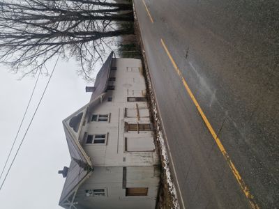 Fotografi av en hvit, to- eller treetasjers bygning med etasjehusform, sett fra siden langs en vei. Bygningen ser ut til å være i dårlig stand med utvislede vinduer og dører.