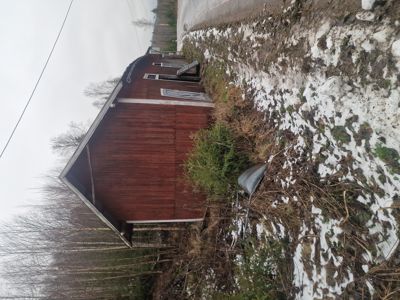 Fotografi av en rødbrun hytte eller uthus sett fra siden, med snø på bakken og skog i bakgrunnen.