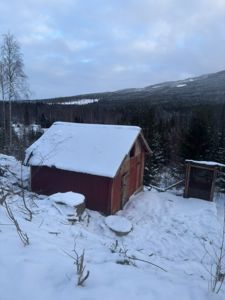 Fotografi av en rød skur eller uthus i snø, sett fra et lavt vinkel med fjell og skog i bakgrunnen.